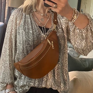 May include: A tan leather bum bag with a gold zip is worn across the body. The bag is paired with a paisley print blouse, black trousers, and silver jewellery. The image is taken in a mirror.