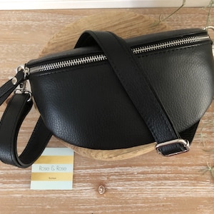 May include: Black leather fanny pack with a silver zipper and adjustable strap. The bag has a curved shape and a small handle on the side. The bag is sitting on a wooden surface.