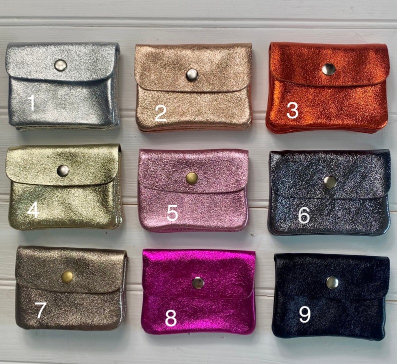May include: Nine small, metallic leather coin purses in various colours: silver, gold, copper, and shades of pink, bronze, and black. Each purse has a flap closure with a single button. Numbers 1-9 are printed on the purses.