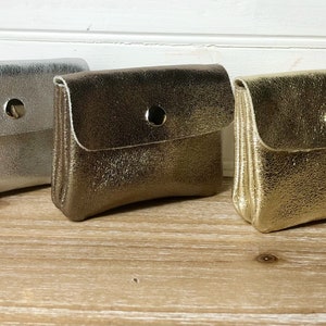 May include: Three small, metallic pouches in silver, bronze, and gold. Each pouch has a flap closure secured by a single snap. The pouches are displayed on a light wooden surface.