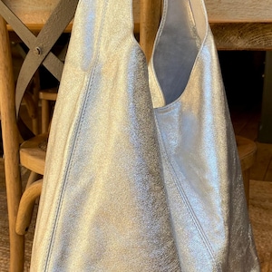 May include: Silver metallic shoulder bag with a large, open design. The bag has a soft, slouchy shape and a single shoulder strap. The material has a textured, crinkled appearance. The bag is displayed on a wooden chair.