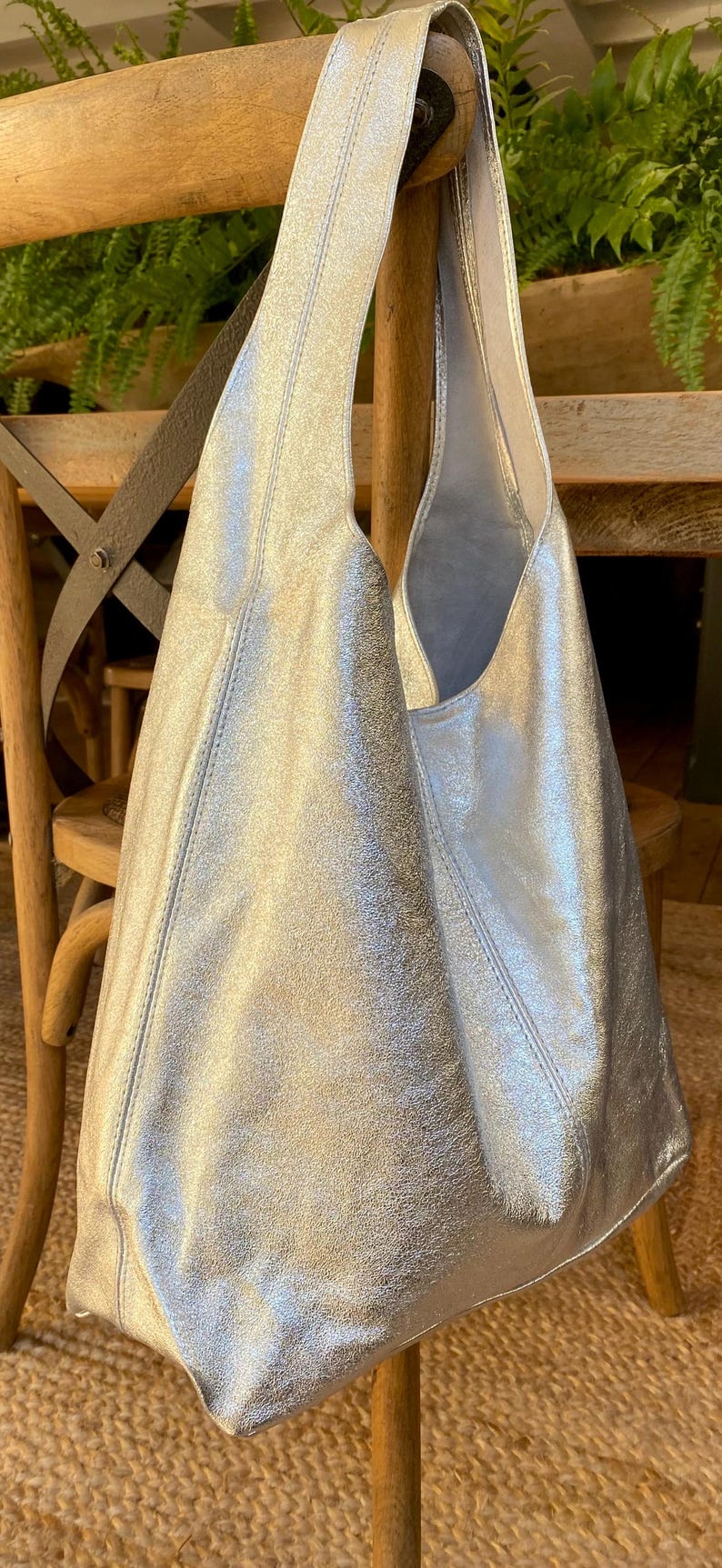 May include: Silver metallic shoulder bag with a large, open design. The bag has a soft, slouchy shape and a single shoulder strap. The material has a textured, crinkled appearance. The bag is displayed on a wooden chair.