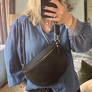 May include: Black leather fanny pack with a gold zipper and hardware. The bag is crescent-shaped and worn across the body. The person is wearing a denim shirt and black jeans.