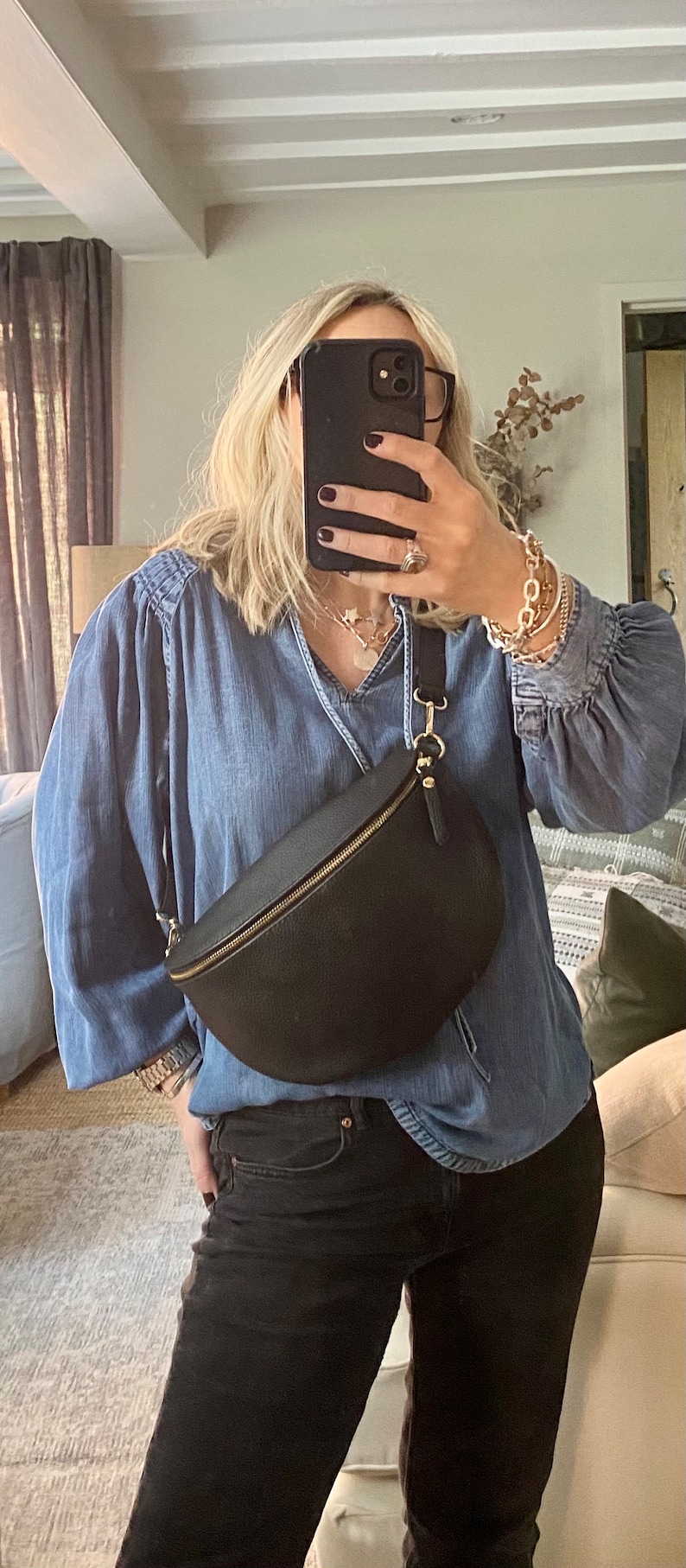 May include: Black leather fanny pack with a gold zipper and hardware. The bag is crescent-shaped and worn across the body. The person is wearing a denim shirt and black jeans.
