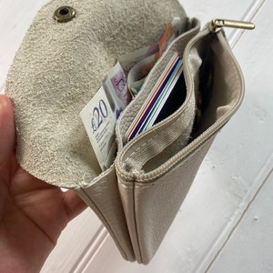 May include: A beige leather wallet with multiple compartments, including a zippered section and a snap closure. The wallet is open, revealing cash, credit cards, and coins. The interior is a lighter shade of beige.