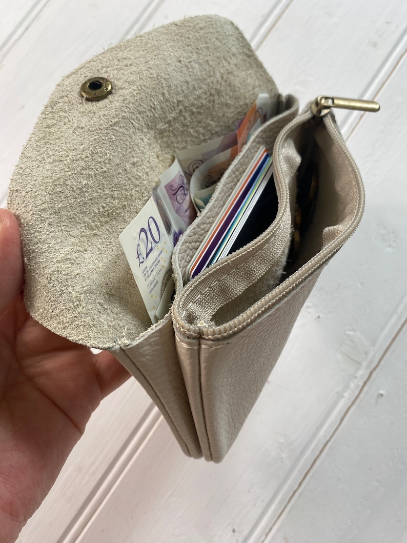 May include: A beige leather wallet with multiple compartments, including a zippered section and a snap closure. The wallet is open, revealing cash, credit cards, and coins. The interior is a lighter shade of beige.