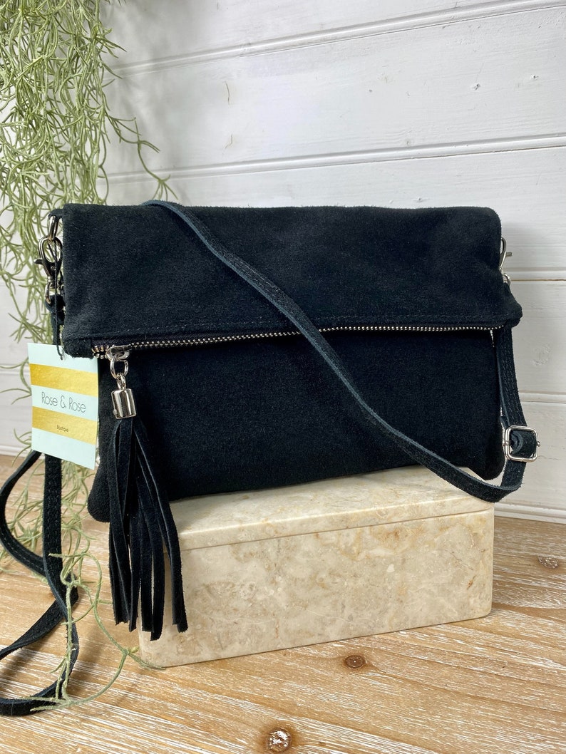May include: Black suede crossbody bag with a silver zipper and tassel. The bag has a long, adjustable strap and a fold-over flap. The tag reads "Rose & Rose Boutique". The bag is displayed on a marble-like box.