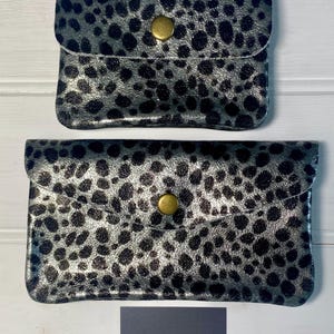 May include: Two silver and black spotted clutch wallets with a flap closure and a gold-coloured button. The wallets are rectangular and have a shiny finish. The bottom of the image shows a logo for Rosearose Boutique.