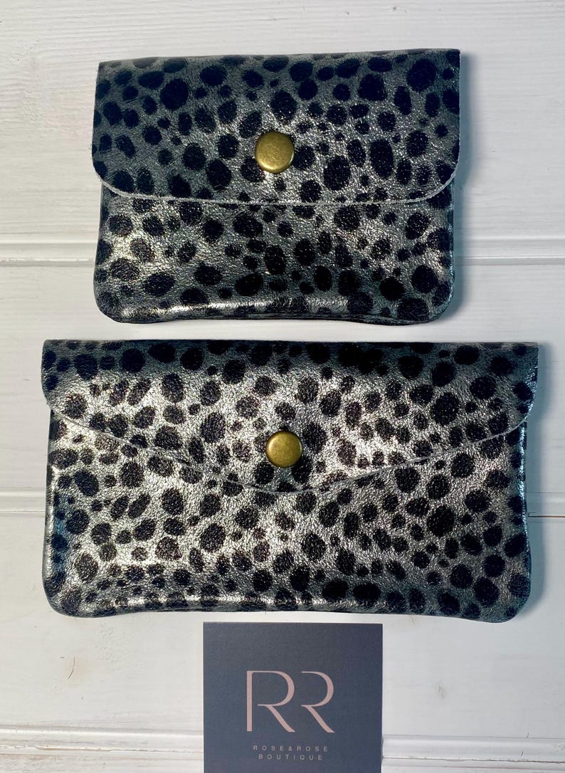 May include: Two silver and black spotted clutch wallets with a flap closure and a gold-coloured button. The wallets are rectangular and have a shiny finish. The bottom of the image shows a logo for Rosearose Boutique.