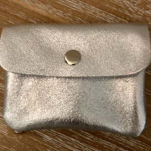 May include: A small, silver pouch with a textured finish. The pouch features a flap closure fastened with a gold-coloured press stud. The rectangular pouch is likely made from leather or a similar material.