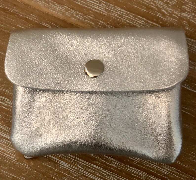 May include: A small, silver pouch with a textured finish. The pouch features a flap closure fastened with a gold-coloured press stud. The rectangular pouch is likely made from leather or a similar material.