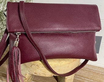 May include: A burgundy leather crossbody bag with a flap closure and a silver zipper. The bag features a matching tassel and a long, adjustable strap. The bag is displayed on a wooden surface.