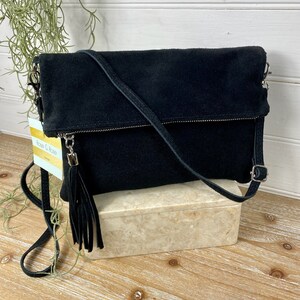 May include: A black suede crossbody bag with a fold-over flap and a long, adjustable strap. The bag features a silver zipper and a tassel. The bag is sitting on a marble-like box.