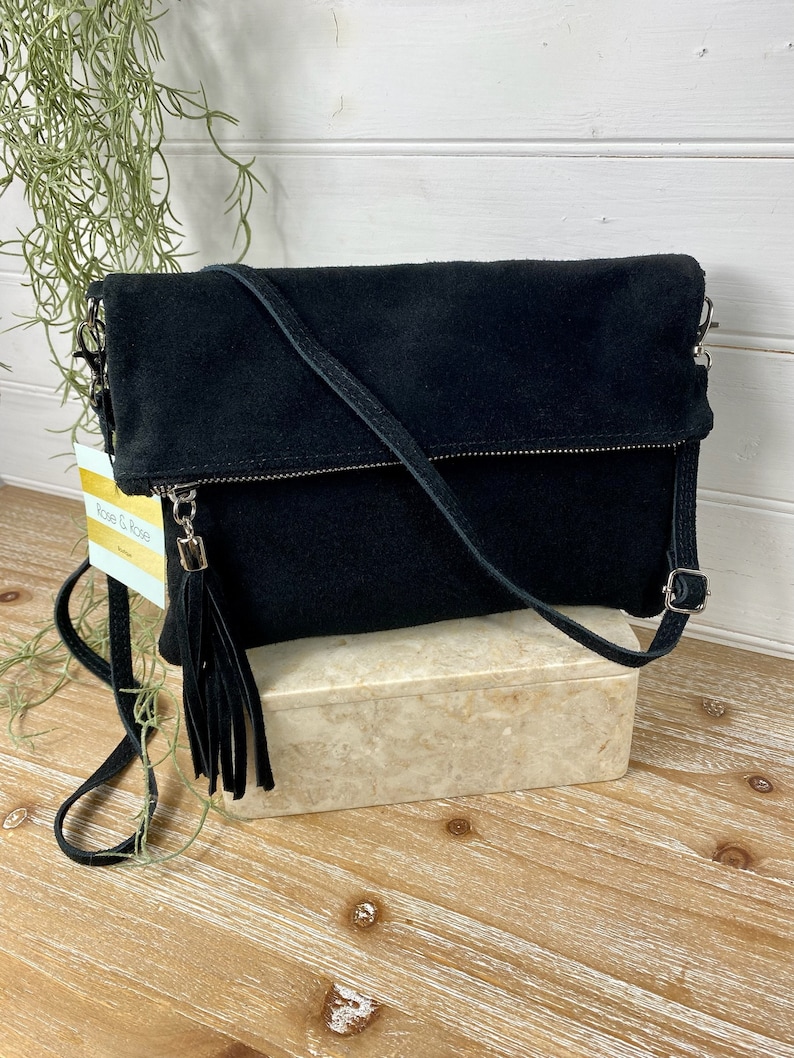 May include: A black suede crossbody bag with a fold-over flap and a long, adjustable strap. The bag features a silver zipper and a tassel. The bag is sitting on a marble-like box.