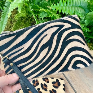 May include: A zebra-print clutch bag with a black leather wrist strap and gold-coloured hardware. The bag has a zip closure and a natural, textured pattern. Green foliage is in the background.