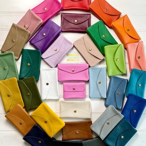 May include: A collection of colorful leather wallets in various shades, including red, pink, green, and blue. Each wallet features a flap closure with a gold-tone button. The wallets are arranged in a circular pattern.