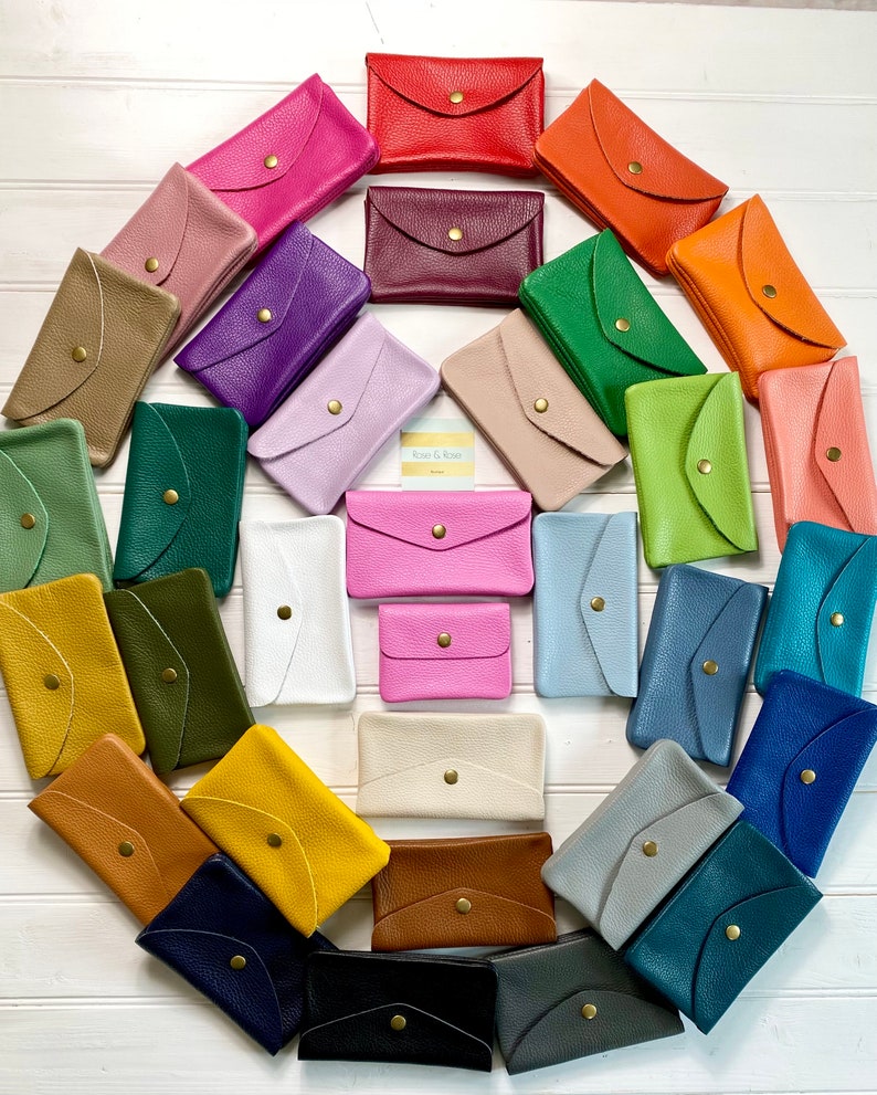 May include: A collection of colorful leather wallets in various shades, including red, pink, green, and blue. Each wallet features a flap closure with a gold-tone button. The wallets are arranged in a circular pattern.