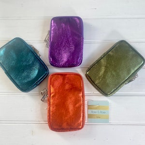 May include: Four metallic zippered pouches in teal, purple, orange, and olive green are displayed on a white wooden surface. Each pouch has a rectangular shape with a silver zipper. A tag with the text "Rose & Rose Boutique" is also present.