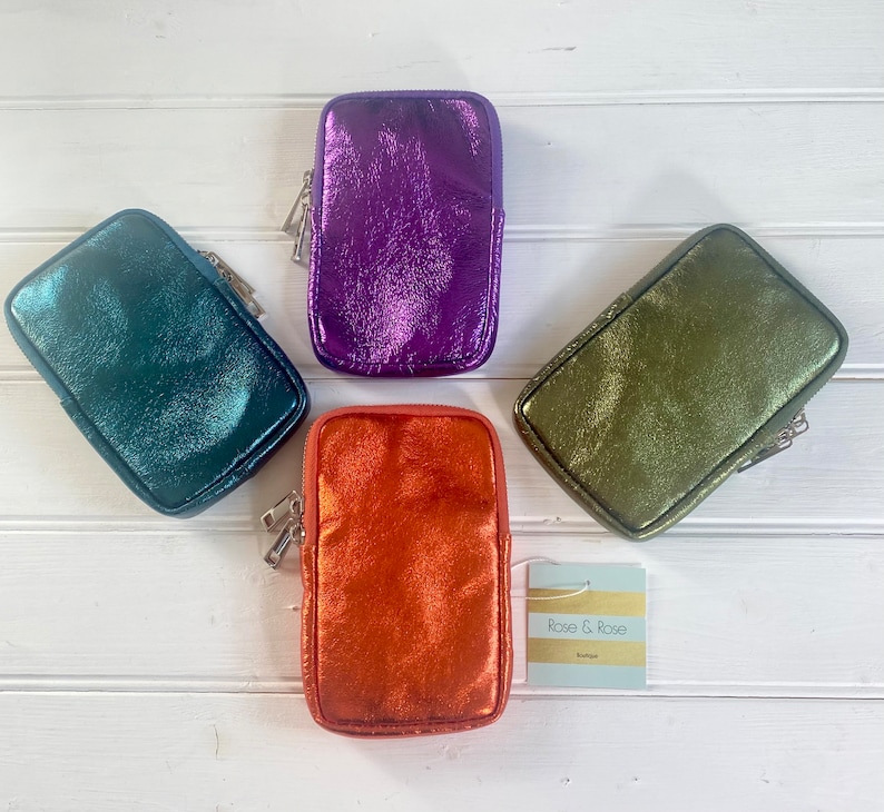 May include: Four metallic zippered pouches in teal, purple, orange, and olive green are displayed on a white wooden surface. Each pouch has a rectangular shape with a silver zipper. A tag with the text "Rose & Rose Boutique" is also present.