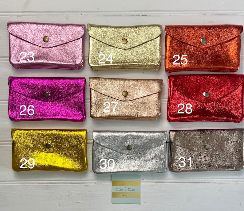 May include: A selection of small, metallic wallets in shades like pink, gold, orange, fuchsia, and silver. Each wallet features a flap closure with a single button. Numbers 23 to 31 are printed on the wallets.
