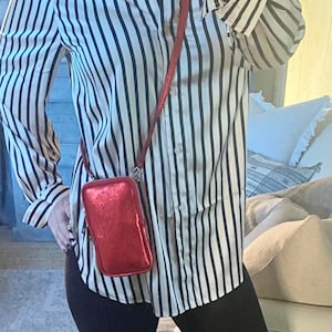 May include: A red, rectangular crossbody phone bag with a zipper closure and a long strap. The bag is metallic and shiny. The person is wearing a black and white striped button-down shirt.