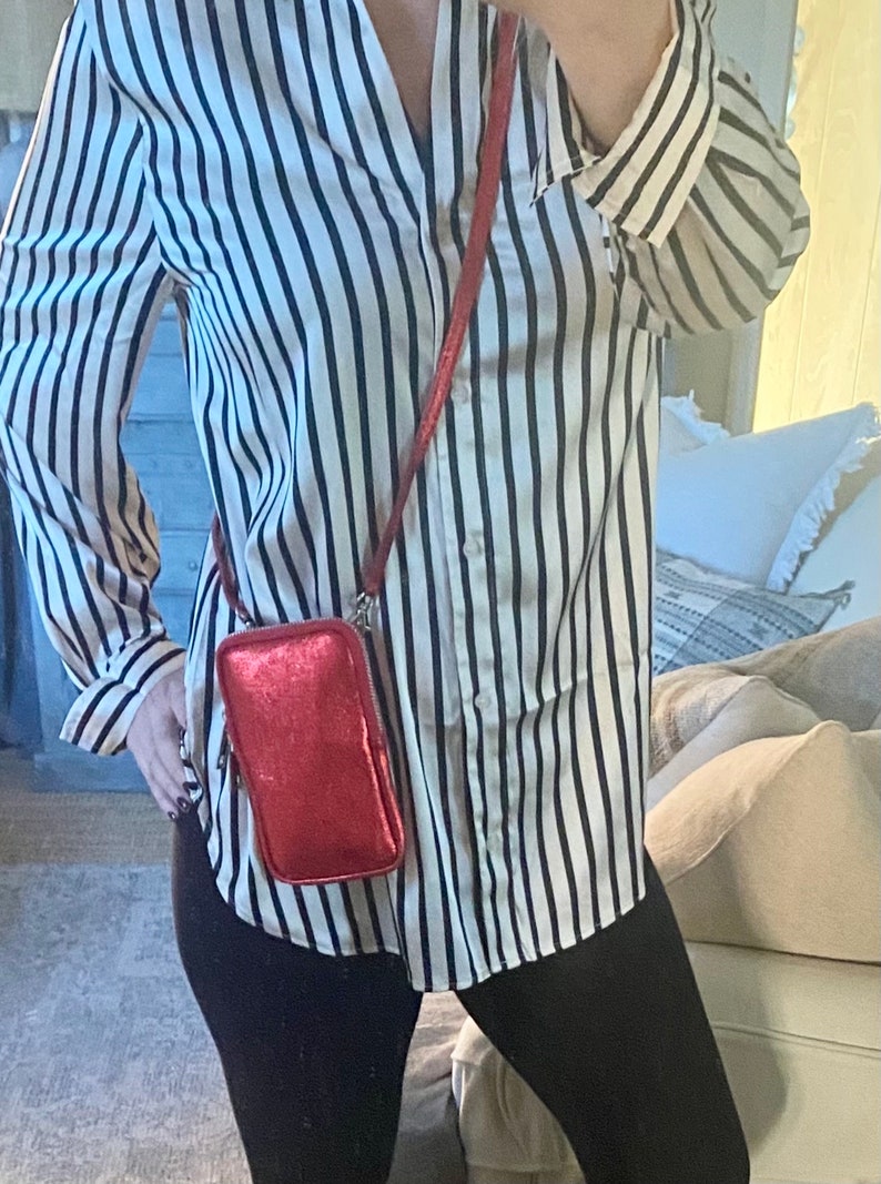 May include: A red, rectangular crossbody phone bag with a zipper closure and a long strap. The bag is metallic and shiny. The person is wearing a black and white striped button-down shirt.