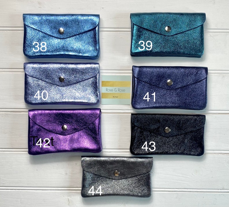 May include: A selection of small, metallic wallets in shades of blue, teal, purple, and black. Each wallet features a flap closure with a silver press stud. The wallets are displayed on a white wooden surface. Numbers 38, 39, 40, 41, 42, 43, and 44 are printed on the wallets.