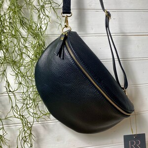 May include: A navy blue, crescent-shaped crossbody bag with a gold zipper and hardware. The bag has a textured surface and a long, adjustable strap. A small tag with the letters "RR" hangs from the bag.