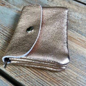 May include: A small, rose gold wallet with a textured finish. The wallet has a flap closure secured by a dark green button. The interior is lined with white stitching. The wallet is closed and appears to be empty.