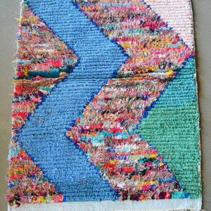 Puede incluir: Una alfombra colorida tejida a mano con un patrón de chevron en tonos de azul, verde, rosa y amarillo. La alfombra tiene un borde de flecos y está hecha de materiales reciclados.