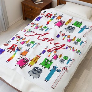 Könnte beinhalten: Weiße Decke mit bunten Cartoon-Zahlenfiguren und dem Namen "Amy" in roter Schrift. Die Decke hat verschiedene Zahlen und Figuren, die ein verspieltes Design schaffen. Die Decke liegt auf einem Bett.