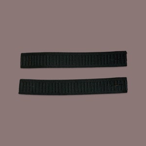 May include: Two black elastic bands, each about 1 inch wide, are shown against a light brown background.