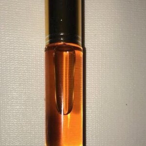 May include: A small, clear glass roll-on bottle filled with a golden-coloured liquid. The bottle has a black cap and a clear glass rollerball. The liquid appears to be oil-based. The bottle is standing upright against a neutral background.