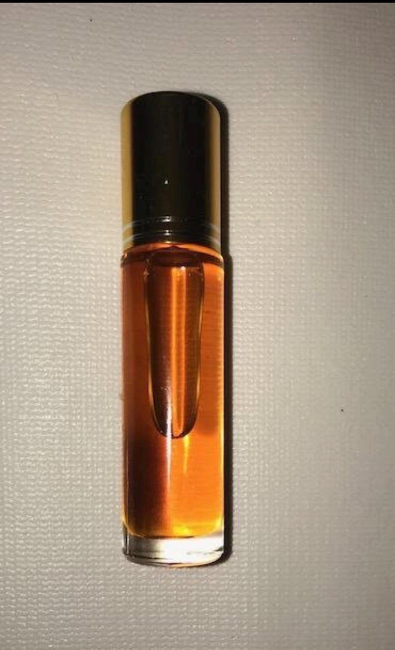 May include: A small, clear glass roll-on bottle filled with a golden-coloured liquid. The bottle has a black cap and a clear glass rollerball. The liquid appears to be oil-based. The bottle is standing upright against a neutral background.
