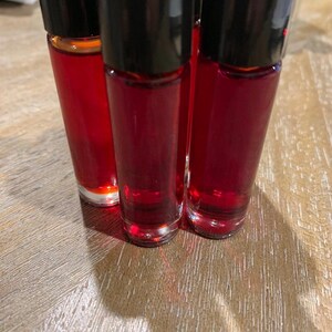 May include: Three small glass bottles filled with a deep red liquid, each with a black cap. The bottles are close together, with the liquid visible through the clear glass. They are on a light brown wooden surface.
