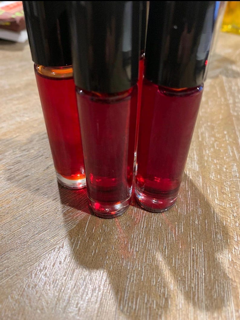 May include: Three small glass bottles filled with a deep red liquid, each with a black cap. The bottles are close together, with the liquid visible through the clear glass. They are on a light brown wooden surface.