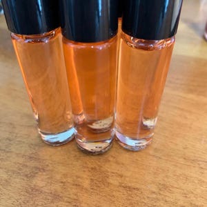 May include: Three clear glass roll-on bottles filled with orange liquid, with black caps. The bottles are arranged on a wooden surface. The bottles are partially in focus, with the liquid inside appearing bright. The bottles are approximately 10 cm tall.