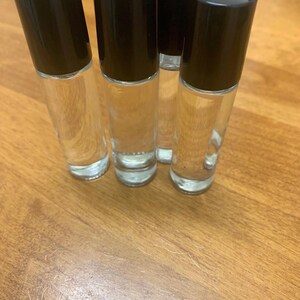May include: Five clear glass roll-on bottles with black caps. The bottles are filled with a clear liquid and are arranged on a wooden surface. The caps are cylindrical and opaque, contrasting with the transparent bottles.