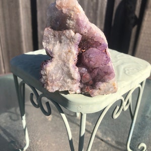May include: A large amethyst crystal cluster with a purple and white color scheme. The crystal is sitting on a small, light green metal stand.
