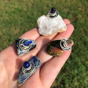 May include: Four small stones with silver crescent moon charms and gemstone tops. The stones are a white crystal, a dark gray stone with white flecks, a green and red stone, and a blue stone.