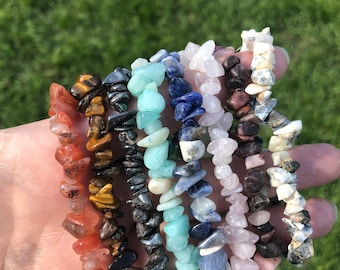May include: A set of eight gemstone bracelets, each featuring a different color and type of stone. The bracelets are made of small, irregular-shaped stones and are stacked on top of each other. The colors of the stones include red, orange, yellow, green, blue, purple, pink, and white.