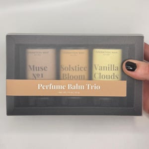 May include: A rectangular black box containing a perfume balm trio with three cylindrical containers labeled Muse No1, Solstice Bloom, and Vanilla Clouds. A tan paper band across the center reads Perfume Balm Trio and Net Wt. 1.5 oz / 42 g.