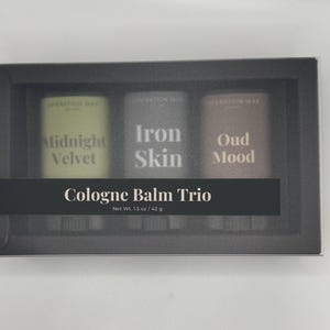 Puede incluir: Un set Cologne Balm Trio en una caja rectangular negra que contiene tres barras de bálsamo. Las etiquetas dicen Midnight Velvet, Iron Skin y Oud Mood. Una banda negra muestra el texto Cologne Balm Trio y Net Wt. 1.5 oz / 42 g.
