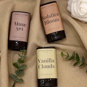 Op de afbeelding: Drie parfumbalmsticks met zwarte bases en bruine etiketten. De etiketten lezen "Muse No. 1", "Solstice Bloom" en "Vanilla Clouds". Elke stick is gelabeld met "Perfume Balm" en "0,5 oz / 14 g". De achtergrond is een bruine stof.
