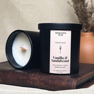 May include: Two black cylindrical candles with white wax and a wooden wick. The label reads Operation Wax, Wood Wick, Vanilla & Sandalwood, Palo Santo, Musk, Sandalwood, Soy-Paraffin Candle, 9 oz / 255 g, with a graphic of a lit match.