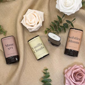 Puede incluir: Vista cenital de tres envases de bálsamo perfumado y un bálsamo suelto. Las etiquetas dicen "Muse No. 1", "Vanilla Clouds" y "Solstice Bloom". El fondo es tela beige, con rosas artificiales.