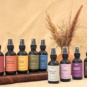Op de afbeelding: Tien matzwarte glazen sprayflessen met het label Operation Wax, elk voorzien van een uniek gekleurd etiket met geurnamen zoals Dark Rose, Amber & Rosewood, Clean Citrus, Cypress Meditation, Salted Orchid, White Tea & Aloe, Hippie, Wisconsin Wildberry, Cashmere & Coconut en Soft Wool.