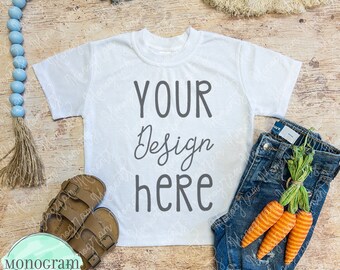 May include: A white t-shirt with the text "YOUR Design here" is displayed on a wooden surface, alongside denim shorts, brown sandals, and decorative carrots. A string of blue beads and a logo are also present.