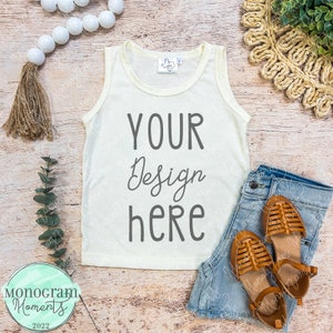 May include: A white tank top with the text "YOUR Design here" on a wooden background. The tank top is lying flat with a pair of denim shorts and brown sandals beside it.