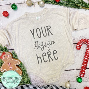 May include: A beige baby onesie with the text "YOUR Design here" printed on the front. The onesie is surrounded by Christmas decorations, including a gingerbread man, candy canes, and a green garland.